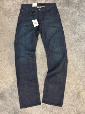 Mörkblå raka jeans från Tiger of Sweden - Snygga mörkblå jeans från Tiger of Sweden med rak passform och klassisk femficksdesign. Jeansen är tillverkade i kraftigt denimtyg och har metallknappar samt nitar. Perfekta för dig som gillar stilrena och tidlösa jeans.