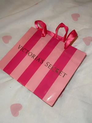 Victoria's Secret shoppingväska rosa - Säljer en ikonisk shoppingväska från Victoria's Secret med rosa och ceriserosa ränder samt glansig finish. Väskan har två rosa satinband som handtag och klassisk logga framtill. Perfekt för dig som gillar snygga accessoarer med statement.