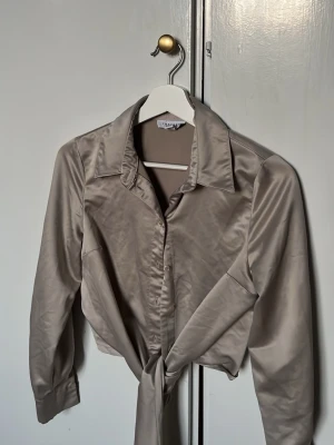 Beige satinblus med knyt framtill - Stilren beige blus i glansigt satinmaterial med klassisk krage och knäppning framtill. Blusen har långa ärmar och en snygg knytning vid midjan som ger en trendig look. Perfekt att matcha med jeans eller kjol för en chic outfit. SÅÅÅ ELEGANTTT
