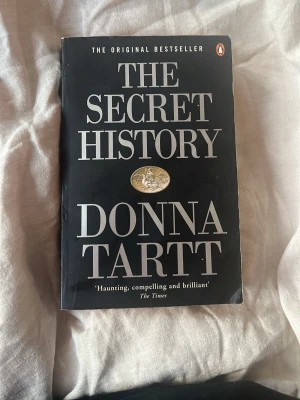 The Secret History av Donna Tartt - En ikonisk roman på engelska, 'The Secret History' av Donna Tartt. Boken har svart mjukt omslag med stora vita bokstäver och ett guldigt emblem på framsidan. Perfekt för dig som gillar spännande och psykologiska berättelser.