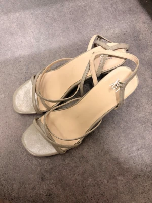 Beige klackade sandaler med remmar - Snygga beige sandaler  med smala remmar och fyrkantig tå. Skorna har en hög blockklack och justerbar rem runt ankeln. Tillverkade i syntetmaterial med en stilren och modern look.