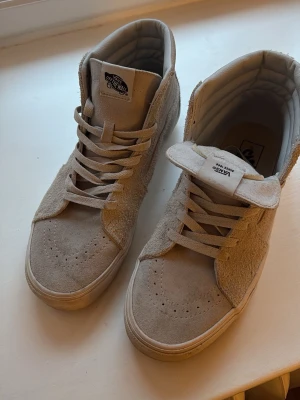 Grå Vans Sk8-Hi sneakers mocka - Snygga beige Vans Sk8-Hi sneakers i mocka med klassisk hög siluett och vadderad krage. Skorna har ton-i-ton snörning, perforerad tå och ikonisk sidestripe. Perfekta för en stilren streetlook.