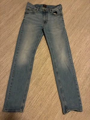 Ljusblå slim fit jeans från Lee - Snygga ljusblå slim fit jeans från Lee med klassisk femficksdesign och diskreta slitningar på låren. Jeansen har normal midja och raka ben, tillverkade i mjuk denim med lite stretch för extra komfort. De är W29 L32