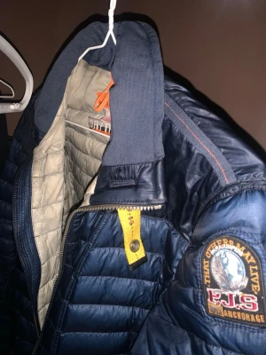 Blå dunjacka från Parajumpers - Snygg blå dunjacka från Parajumpers med quiltad design och emblem på ärmen. Jackan har gul detalj vid dragkedjan och kontrastfärgad insida i beige. Perfekt för kalla vinterdagar och har en sportig look.