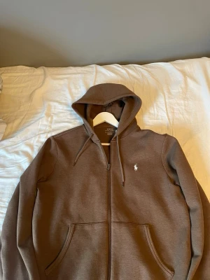 Brun hoodie från Polo Ralph Lauren - Nypris över 2000 kr. Extremt snygg brun hoodie från Polo Ralph Lauren med dragkedja framtill och klassisk huva, tröjan passar alla stilar . Hoodien har fickor på magen och den ikoniska vita logon broderad på bröstet. Perfekt för en chill och stilren look. Tillverkad i mjukt material för extra komfort. Hör av dig med funderingar eller prisförslag!
