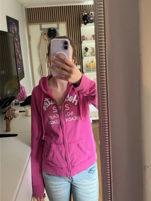 Rosa hoodie från Hollister - Säljer en rosa hoodie från Hollister med dragkedja och vit text på bröstet. Hoodien har huva med dragsko och två fickor framtill. Perfekt för en avslappnad och chill stil. Storlek M men passar på mig som har S. Den har ett litet hål som man kan se på bilden😊
