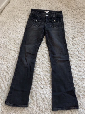 Svarta bootcut jeans med fickdetaljer - Snygga svarta bootcut jeans med två framfickor med lock och knappar. Jeansen har klassiska bakfickor och subtila slitningar för en cool look. Tillverkade i stretchigt denim för skön passform.