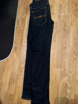 Mörkblå jeans från Nudie Jeans - Snygga mörkblå jeans från Nudie Jeans med klassisk femficksdesign och kontrastsömmar. Jeansen har rak passform och är tillverkade i ett slitstarkt denimtyg. Perfekta för en stilren och enkel look.
