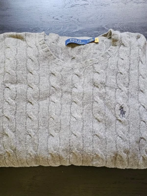 Grå kabelstickad tröja Polo Ralph Lauren - Kabelstickad grå tröja från Polo Ralph Lauren med rund halsringning och broderad logga på bröstet. Tröjan har långa ärmar och är tillverkad i mjukt material som passar perfekt för lager-på-lager-stil. 