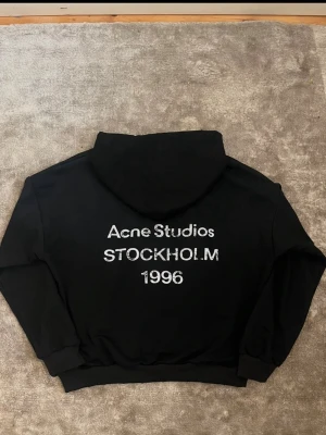 Svart Acne Studios Stockholm hoodie - Svart hoodie från Acne Studios med stor vit text på ryggen: 'Acne Studios STOCKHOLM 1996'. Klassisk passform med huva och ribbade muddar. Tillverkad i mjukt material som känns skönt mot huden. Perfekt för dig som gillar stilrena och trendiga plagg.☺️OBS!!! PRISET KAN DISKUTERAS!!!