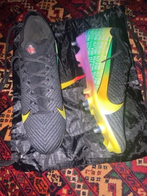 Nike Mercurial färgglada fotbollsskor Vini jr Limited edition Elites SG  - Snygga Nike Mercurial fotbollsskor i svart med gula detaljer och en färgglad sula i regnbågens färger. Skorna har stickad ovandel och klassisk snörning. Perfekta för dig som vill sticka ut på planen med både stil och prestanda.