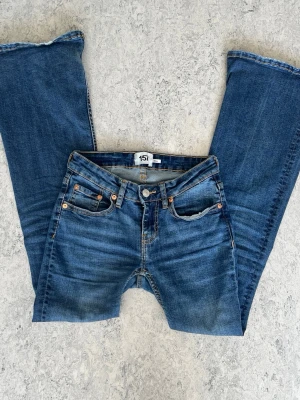 Blå bootcut jeans från 157 - Snygga blå bootcut jeans från 157 med klassisk femficksdesign och ljusa slitningar framtill. Jeansen har normal midja, raka ben och detaljer i kopparfärgad metall. Perfekta för en avslappnad och trendig look.