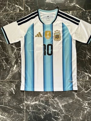 Argentina matchtröja Adidas nr 10 - Säljer en officiell Argentina fotbollströja från Adidas med nummer 10 på bröstet. Tröjan har klassiska blå och vita ränder, svarta detaljer och guldiga FIFA- och AFA-emblem. Tillverkad i lätt och ventilerande polyester, perfekt för fotbollsträning eller match.