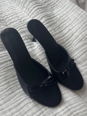 Svarta slip-in pumps med rosett - Stilrena svarta slip-in pumps med öppen tå och liten klack. Skorna har en söt rosettdetalj framtill och är tillverkade i ett slätt material som ger en elegant look. Perfekta för dig som gillar klassisk och enkel stil.
