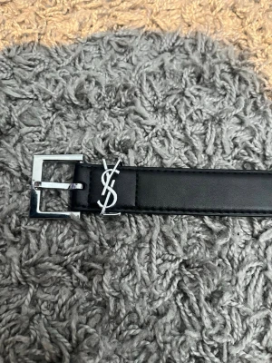 Svart YSL bälte i skinn - Stilrent svart bälte från Yves Saint Laurent i skinn med silverfärgat YSL-logo och spänne. Perfekt accessoar för att lyfta din outfit med en lyxig touch och klassisk design.