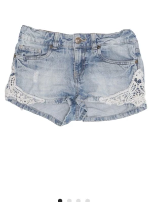 Ljusblå jeansshorts med spetsdetaljer - Snygga ljusblå jeansshorts med vita spetsdetaljer på sidorna. Klassisk femficksmodell med knapp och dragkedja framtill. Perfekta för varma dagar och ger en cool, avslappnad vibe.
