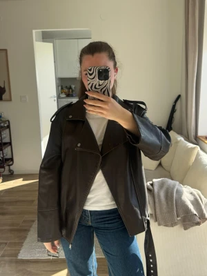 Svart biker skinnjacka med bälte - Cool svart skinnjacka i klassisk bikerstil med sned dragkedja, axelklaffar och bälte i midjan. Jackan har breda slag och silverfärgade detaljer som ger en edgy look. Perfekt för dig som vill ha en tidlös och trendig jacka till din garderob.