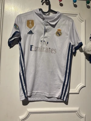 Real Madrid Ronaldo matchtröja - Säljer en vit Real Madrid matchtröja från Adidas med blå detaljer och klubbmärke på bröstet. Tröjan har trycket 'Ronaldo 7' på ryggen och FIFA-märke på framsidan. Tillverkad i lätt och ventilerande material, perfekt för fotbollsträning eller match.
