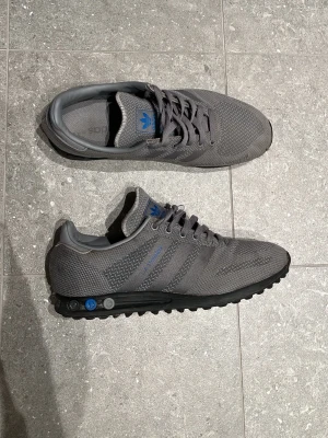 Limiterade Grå Adidas runners  med blå detaljer - Snygga grå runners från Adidas med stickad ovandel och diskreta mönster. Skorna har blå detaljer på plösen och sulan samt klassiska tre ränder på sidan. Yttersulan är svart och har bra grepp. Perfekta för en stilren och sportig look.