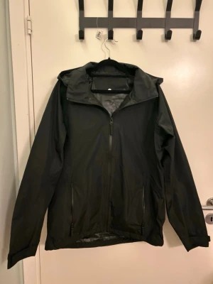 Svart regnjacka med huva - Snygg svart regnjacka med dragkedja framtill och två fickor med dragkedja. Jackan har en klassisk passform och justerbar huva, perfekt för regniga dagar. Tillverkad i vattenavvisande material som håller dig torr när det ösregnar.