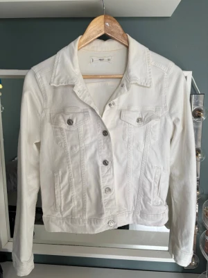 Vit jeansjacka mango - Snygg vit jeansjacka med klassisk krage och silvriga knappar framtill. Jackan har två bröstfickor med lock och knapp samt två sidofickor. Perfekt att slänga över en t-shirt för en fräsch look.