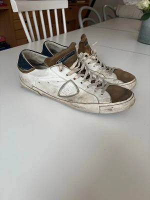 Philippe Model vita och bruna sneakers - Säljer ett par Philippe Model sneakers i vitt och brunt med blå detaljer på hälen. Skorna har mockadetaljer på tån och plösen, samt klassisk logga på sidan. Låga i modellen och med vita snören. Perfekta för dig som gillar en avslappnad och trendig stil. Skorna är använda och slitna på framförallt insida sula. Som skulle kunna bytas ut, men annars är varan i ett okej skick! Pris går att förhandlas!