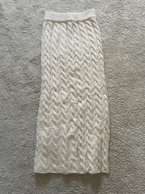 Beige stickad långkjol med slits - Supermysig beige långkjol i stickat material med grovt kabelstickat mönster. Kjolen har en liten slits på sidan och resår i midjan för extra komfort. Perfekt för dig som vill ha en chill och snygg look under kallare dagar.