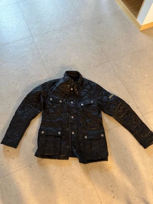 Svart quiltad jacka från Barbour - Ariel Quilted Jacket Black, med klassisk krage, tryckknappar och flera fickor framtill. Jackan har lång ärm och detaljer i silver. Perfekt för dig som vill ha en stilren och tidlös look. 