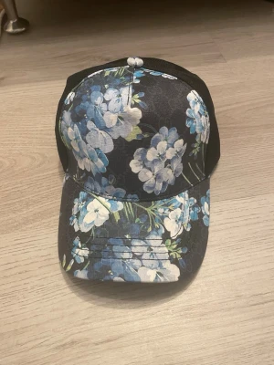 Svart keps med blåblommigt mönster - Snygg svart keps med blommigt mönster i blått och vitt. Framsidan och skärmen är täckta av blommor medan baksidan är i svart mesh för extra ventilation. Perfekt accessoar för en trendig och avslappnad stil.