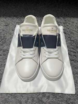 Valentino Garavani vita sneakers - Snygga vita sneakers från Valentino Garavani med en bred mörkblå remsa över sidan och tungan. Skorna har vita snören, guldlogga på plösen och detaljer i läder. Sulan är vit med diskret mönster för extra grepp. Perfekta för dig som gillar exklusiv och stilren design.