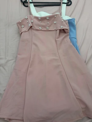 Rosa kort klänning med pärldetaljer - Söt rosa kort klänning med smala axelband och pärldetaljer upptill. Klänningen har en lätt utsvängd form och är perfekt för dig som gillar en feminin och trendig stil.