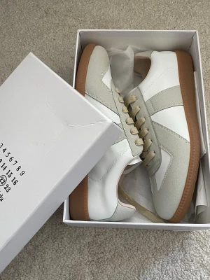 Maison Margiela sneakers  - Snygga Maison Margiela  sneakers i vitt och ljusgrått läder och mocka med beigea snören och klassisk gummisula. Perfekt för dig som vill ha en stilren och trendig look. Skorna kommer med originalkartong och dustbag.