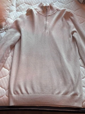 Beige stickad tröja med halvzip - Snygg beige stickad tröja med halv dragkedja och hög krage. Tröjan har lång ärm och ribbade muddar vid ärmslut och nederkant.