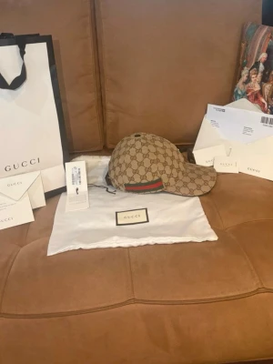 Gucci keps med GG-mönster - Snygg beige keps från Gucci med klassiskt GG-monogram över hela kepsen och en grön-röd rand på sidan. Kepsen har böjd skärm och justerbar rem baktill. Tillverkad i canvasmaterial med exklusiv känsla.