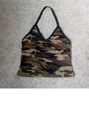 Camo halterneck linnetopp - Snygg camouflagemönstrad halterneck linnetopp med v-ringning och svarta kanter. Perfekt för dig som vill sticka ut med en edgy stil. Toppens material känns mjukt och stretchigt, vilket gör den bekväm att bära.