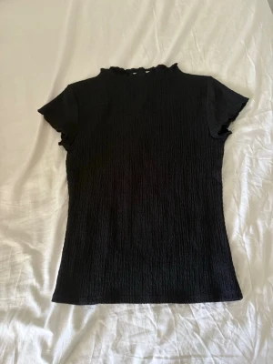 Svart topp med rynkad struktur Kappahl - Svart kortärmad topp från Kappahl i storlek XS. Toppen har en rynkad, stretchig struktur och liten volangkant vid halsringningen. Materialet är mjukt och följsamt, perfekt för en stilren look.