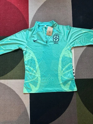 Nike Brasil långärmad träningströja - Snygg långärmad träningströja från Nike med Brasil-logga. Tröjan är turkos med ljusgröna mönster och har halv dragkedja framtill. Perfekt för fotbollsträning eller annan sport. Materialet är lätt och andas bra.