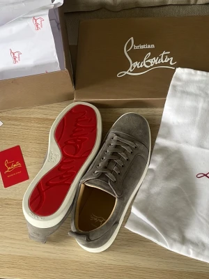 Gråa sneakers från Christian Louboutin - Snygga gråa sneakers från Christian Louboutin med klassisk röd sula och vita kanter. Skorna är tillverkade i mocka och har en låg profil med snörning. Perfekt för dig som vill ha en exklusiv och stilren look. Storlek 42 och 43 finns. HELT NYA OANVÄNDA 10/10 skick. Pris kan diskuteras 