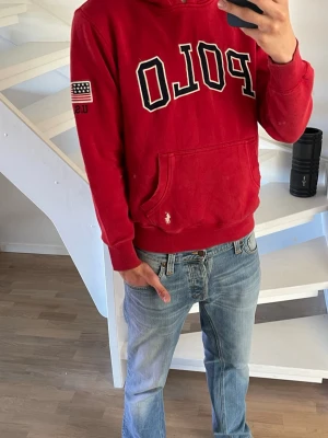 Röd hoodie från Polo Ralph Lauren - Röd hoodie från Polo Ralph Lauren med stor POLO-logga på bröstet, amerikansk flagga och 'USA' på ärmen. Klassisk känguruficka framtill och broderad logga nedtill. Tröjan passar inte som XL utan mer som L