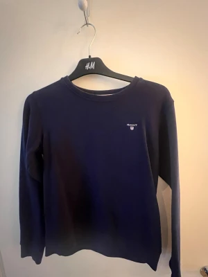 Mörkblå långärmad tröja från GANT - Mörkblå långärmad tröja från GANT med rund halsringning och diskret logga på bröstet. Enkel och stilren design som passar perfekt till jeans eller kjol. Mjuk och bekväm bomullskänsla.