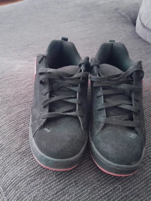 Svarta DC sneakers med rosa detaljer - Säljer ett par svarta DC sneakers med rosa logga och rosa sula. Skorna har klassisk låg profil, snörning och är tillverkade i mockaliknande material. Perfekta för dig som gillar streetwear och vill sticka ut med färg.