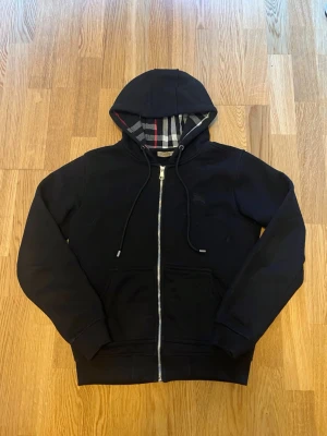 Svart hoodie med dragkedja Burberry - Svart hoodie från Burberry med hel dragkedja och klassiskt rutigt foder i huvan. Tröjan har två stora fickor framtill och justerbar dragsko i huvan. Perfekt för en stilren och avslappnad look.