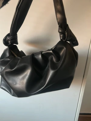 Svart handväska med knutdetaljer - Trendig svart handväska i mjukt fejkläder med coola knutdetaljer på axelremmen. Väskan har en rymlig insida med ljus textilfodring och stängs med dragkedja. Perfekt för dig som gillar en stilren men edgy look.