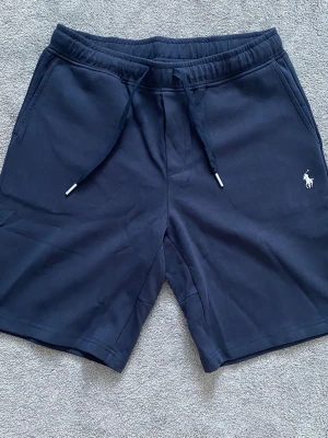 Ralph lauren shorts - Mörkblå shorts från Polo Ralph Lauren med klassisk logga broderad på vänster ben. Modellen har snörning i midjan, två sidofickor och en bakficka. Tillverkade i mjuk bomull för en skön och avslappnad stil. Storlek L men mer som M. Passar om du är 175-185cm. Använt 2 gånger