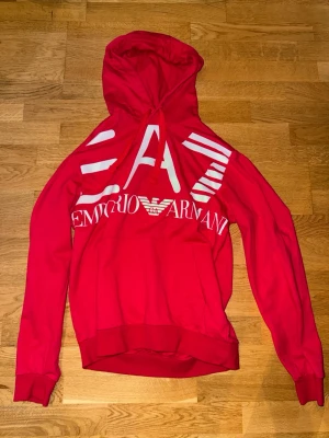 Röd hoodie från Emporio Armani - Snygg röd hoodie från Emporio Armani med stor vit logga och text framtill. Hoodien har huva med dragsko och ribbade muddar vid ärmslut och nederkant. Perfekt för dig som vill sticka ut med en trendig och bekväm look.