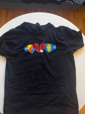 Svart t-shirt med boxningsmotiv - Svart t-shirt med tryck av boxningshandskar mellan två klubbmärken på bröstet. Klassisk passform och rund halsringning. Köpt på sofialäntaren, perfekt för dig som är djurgårdare.