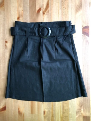 Svart kjol med bälte och ringdetalj - Stilren svart kortkjol med hög midja och brett bälte med stor rund spänne. Kjolen har veck framtill och är lätt utsvängd. Perfekt för dig som gillar en enkel men trendig look.