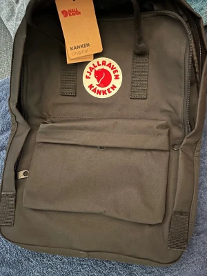 Svart Fjällräven Kånken ryggsäck - Klassisk svart Kånken ryggsäck från Fjällräven med ikonisk röd logotyp framtill. Ryggsäcken har ett stort huvudfack, en framficka med dragkedja och två sidofickor. Tillverkad i slitstarkt syntetmaterial och har bärhandtag upptill. Med sittdyna. Finns ett märke som syns i solen på loggan. Köpes i befintligt skick