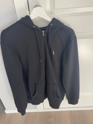 Svart hoodie från Polo Ralph Lauren - Inprincip oanvänd, Svart hoodie med dragkedja från Polo Ralph Lauren. Klassisk design med huva, dragsnören och två fickor framtill. Liten vit logga broderad på bröstet. Skön och stilren, perfekt till jeans eller joggers.