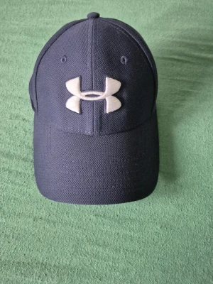 Svart keps från Under Armour - Snygg svart keps från Under Armour med böjd skärm och stort vitt logotryck framtill. Kepsen har klassisk sexpanelsdesign och är tillverkad i ett slitstarkt tyg med ventilerande hål upptill. Perfekt för en sportig och avslappnad stil.